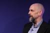 photo of Neal Stephenson, a bald man with a big black and grey goatee, against a blue background. He's wearing a suit jacket, has a lapel microphone on, and appears to be speaking.