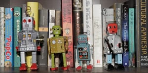 A closeup photo of a shelf of books with a row of four toy robots in front of the books. 